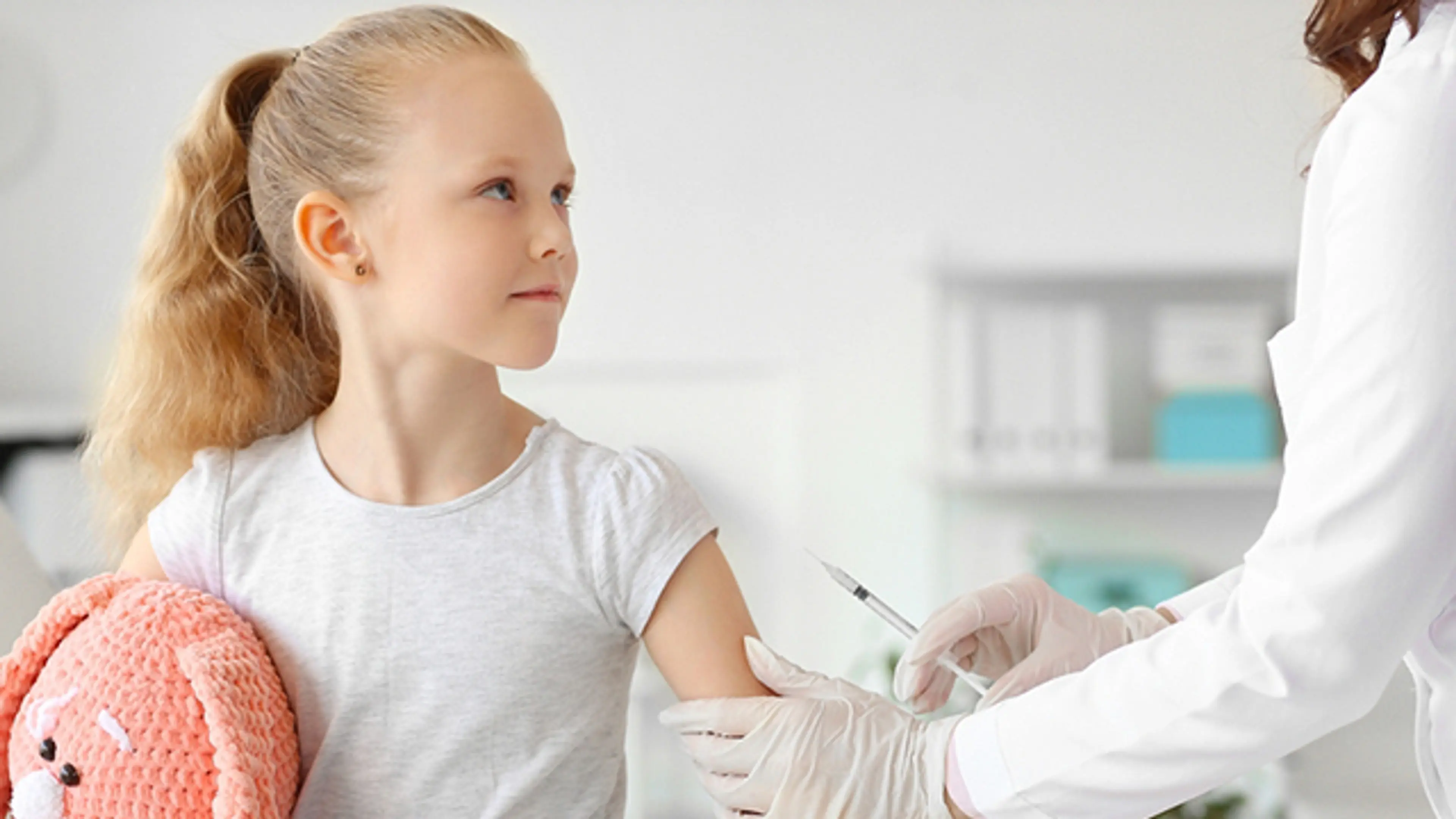 Nurse administering a vaccination injection to a young girl holding a stuffed toy during a pediatric immunization visit.