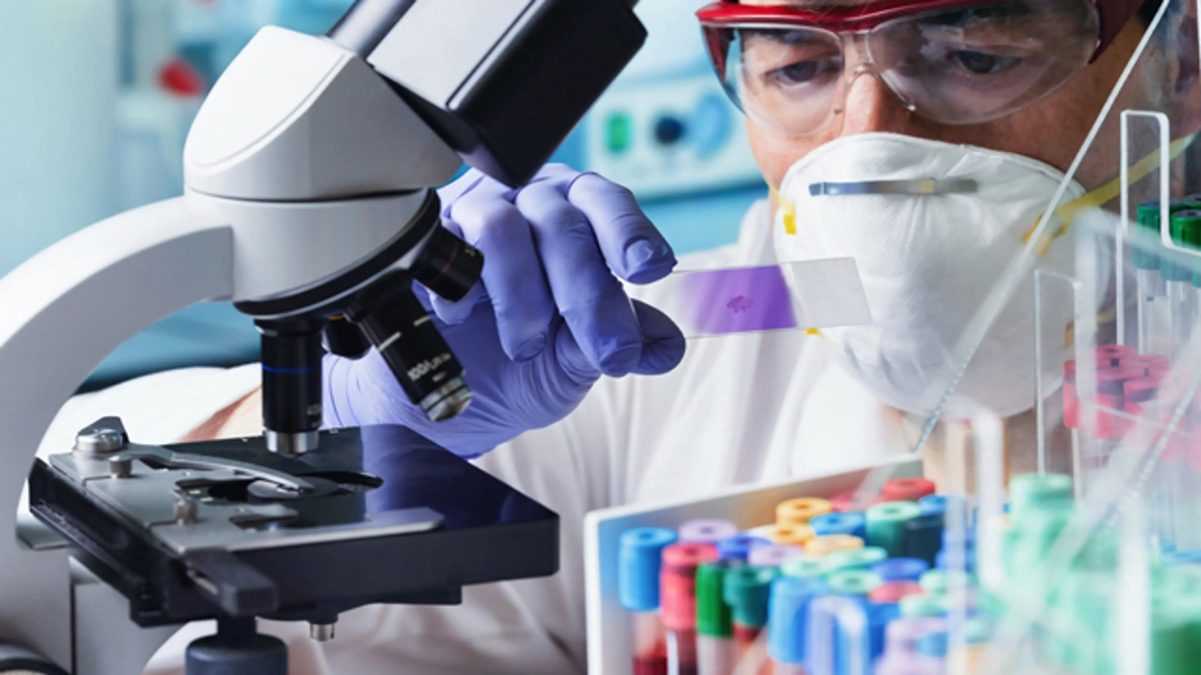 Laboratory technician analyzing a blood sample under a microscope with test tubes in a clinical diagnostic laboratory for blood testing services.