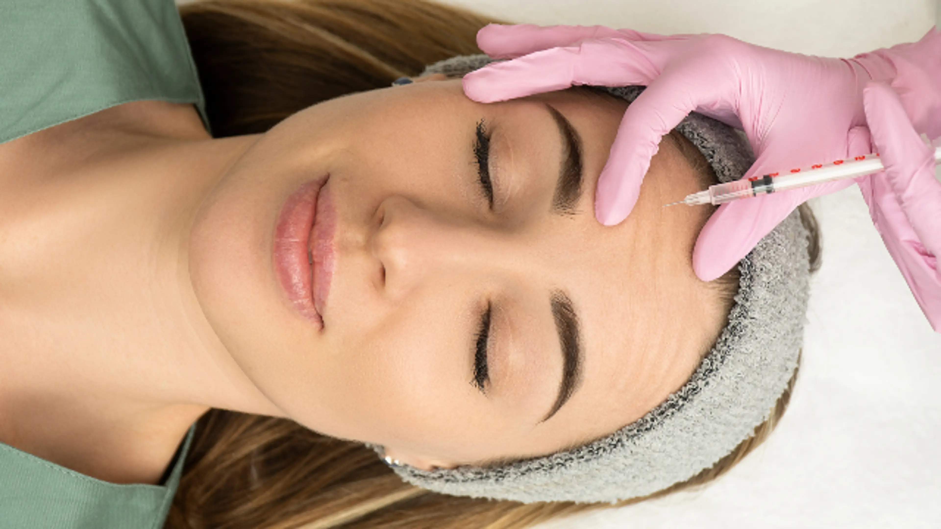 A woman lies with her eyes closed while a practitioner in pink medical gloves administers an injection into her forehead using a fine-tipped syringe.