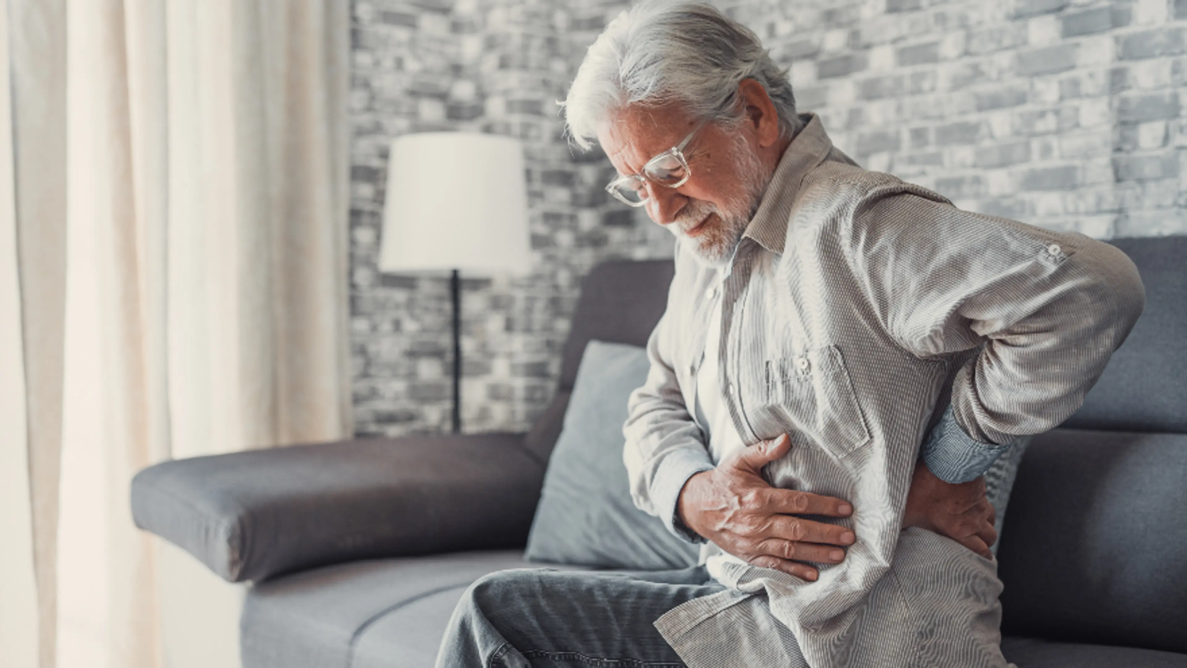 Elderly man sitting on a sofa, holding his lower back in pain