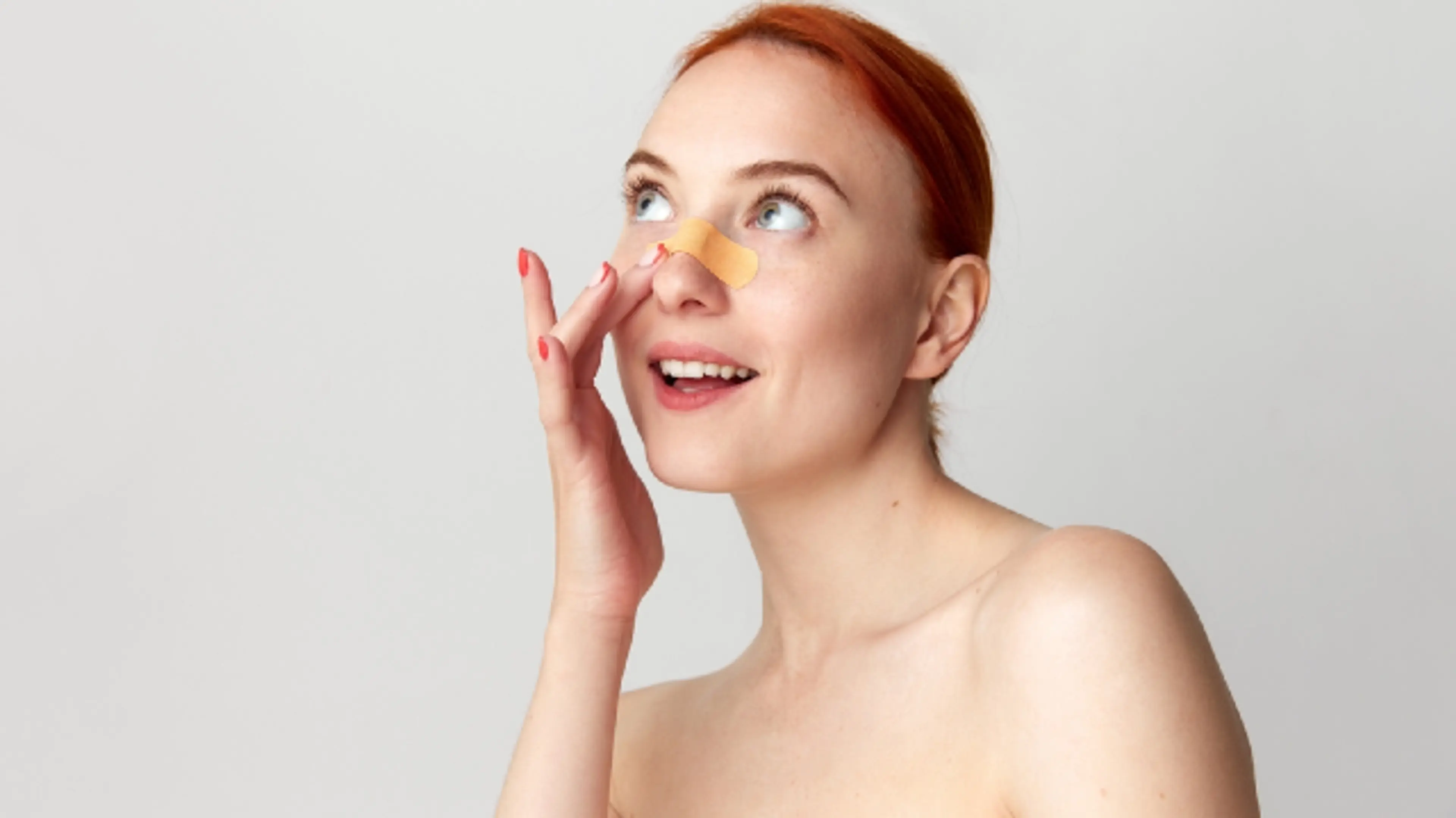A woman with red hair applies or adjusts a small adhesive bandage on her nose while looking upward with a surprised, cheerful expression against a plain light background.
