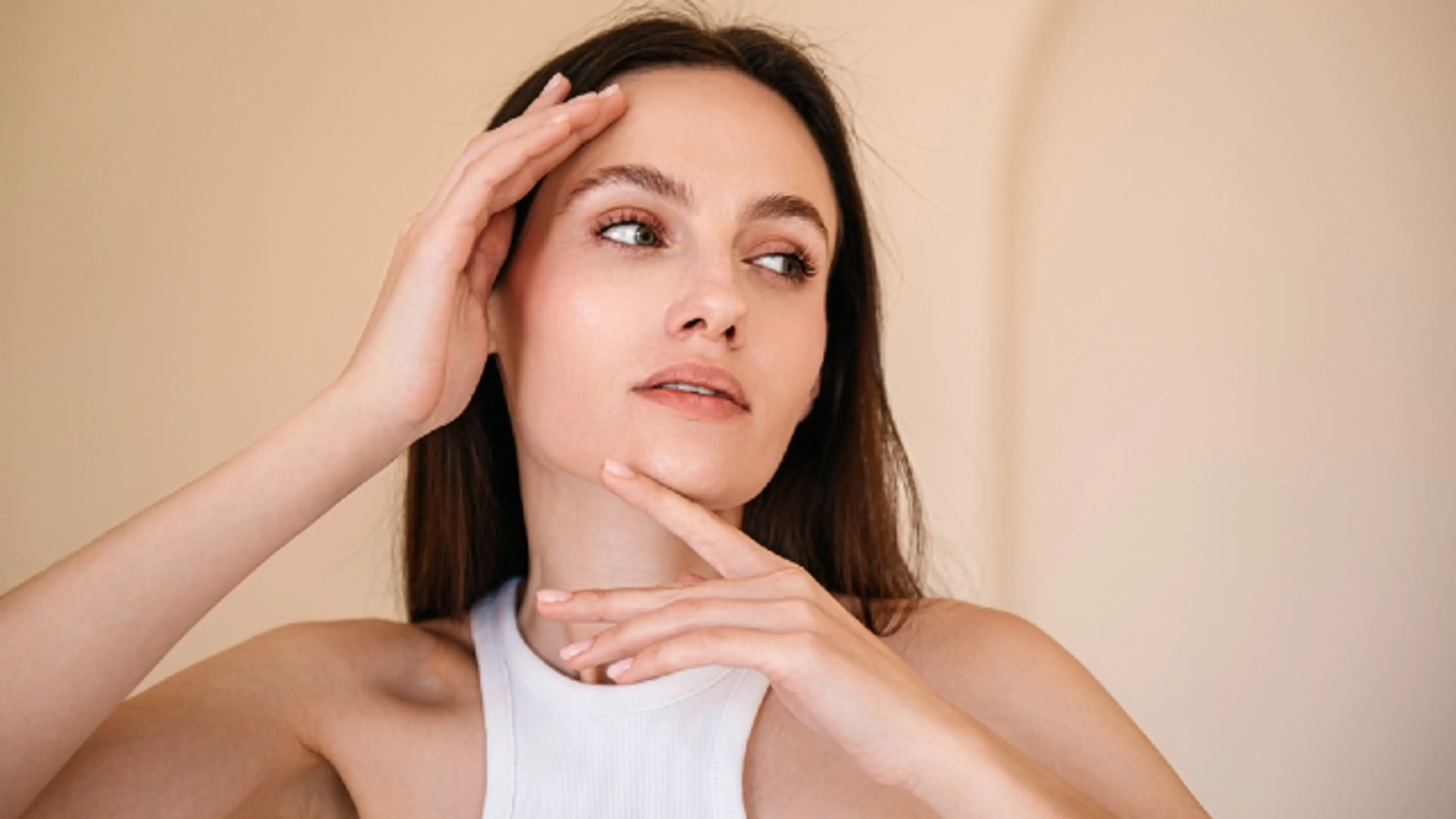 A young woman with clear skin gently touching her face, looking slightly to the side, in a soft beige background—suggesting skincare, beauty, and a natural glow.
