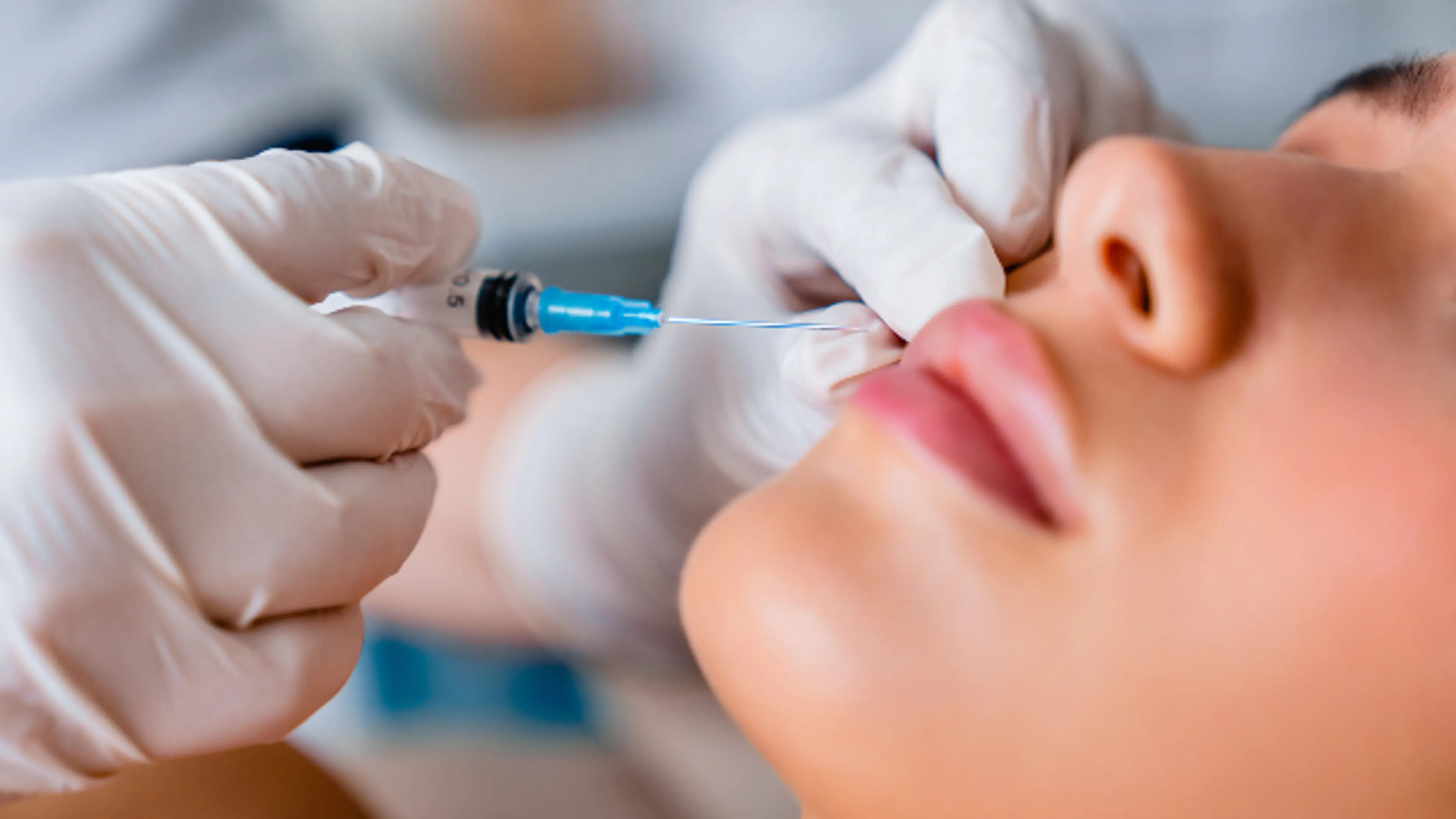 A practitioner in white medical gloves administering a cosmetic injection into the upper lip area of a woman.