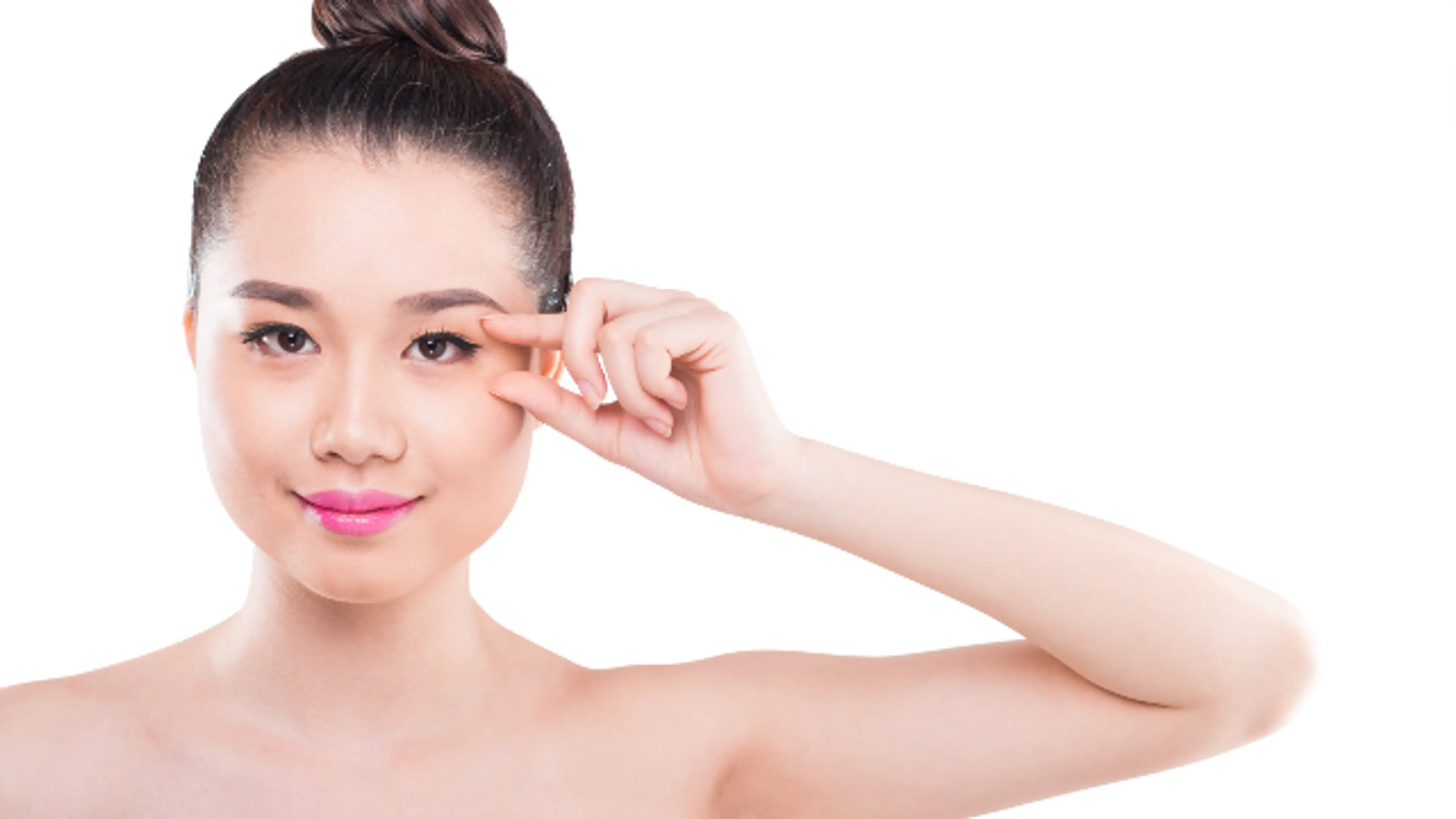 A young woman with her hair in a bun gestures toward her eyelid while looking into the camera against a plain white background.