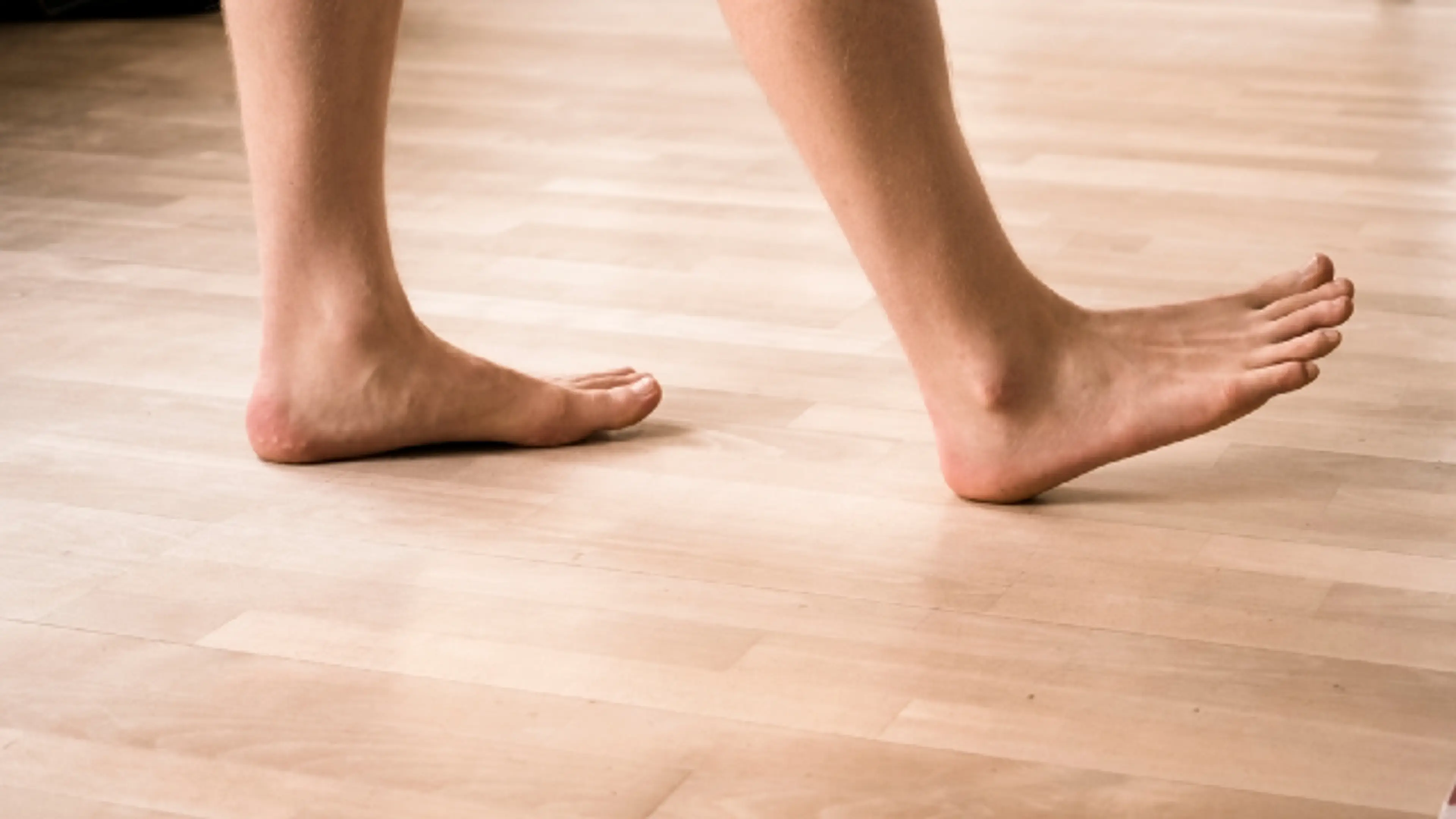 A side-angle, close-up shot of a person’s bare feet walking across a light-colored wooden floor, with the front foot flexed upward mid-stride.