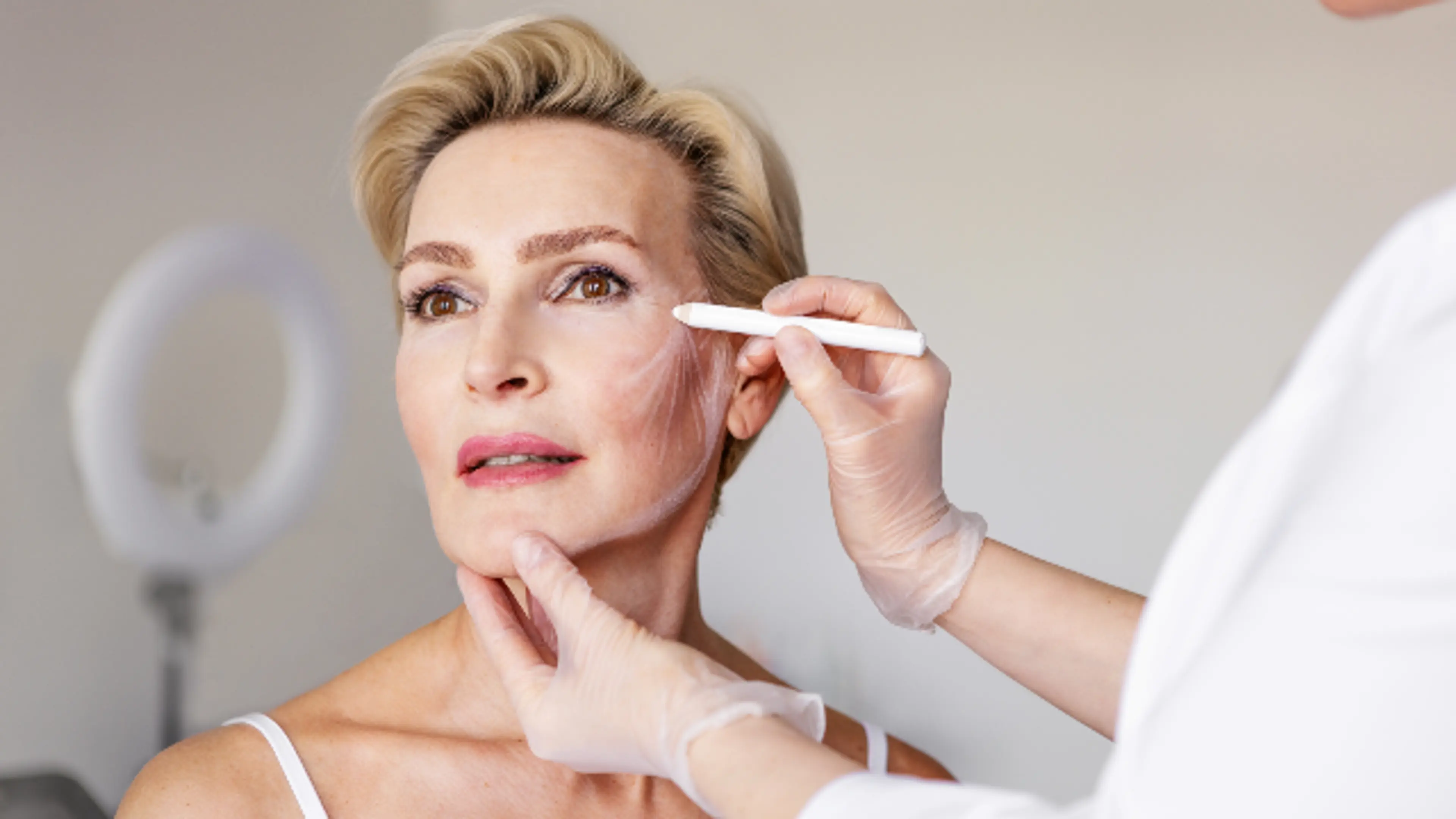 A professional in translucent gloves using a white marking tool to map out treatment lines on the cheek of a mature woman with blonde hair.