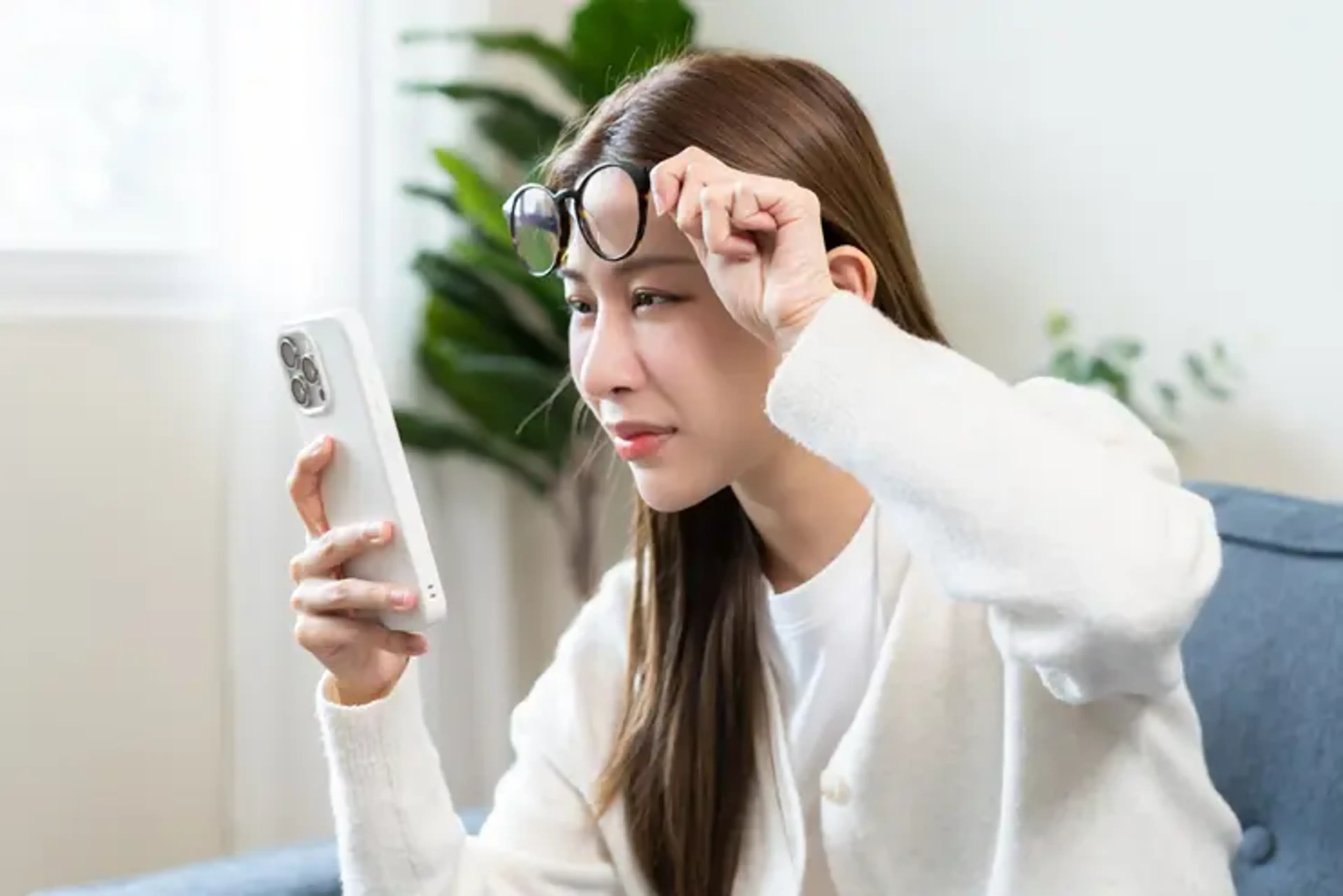 Woman holding a smartphone while lifting her reading glasses, illustrating presbyopia and difficulty with near vision.